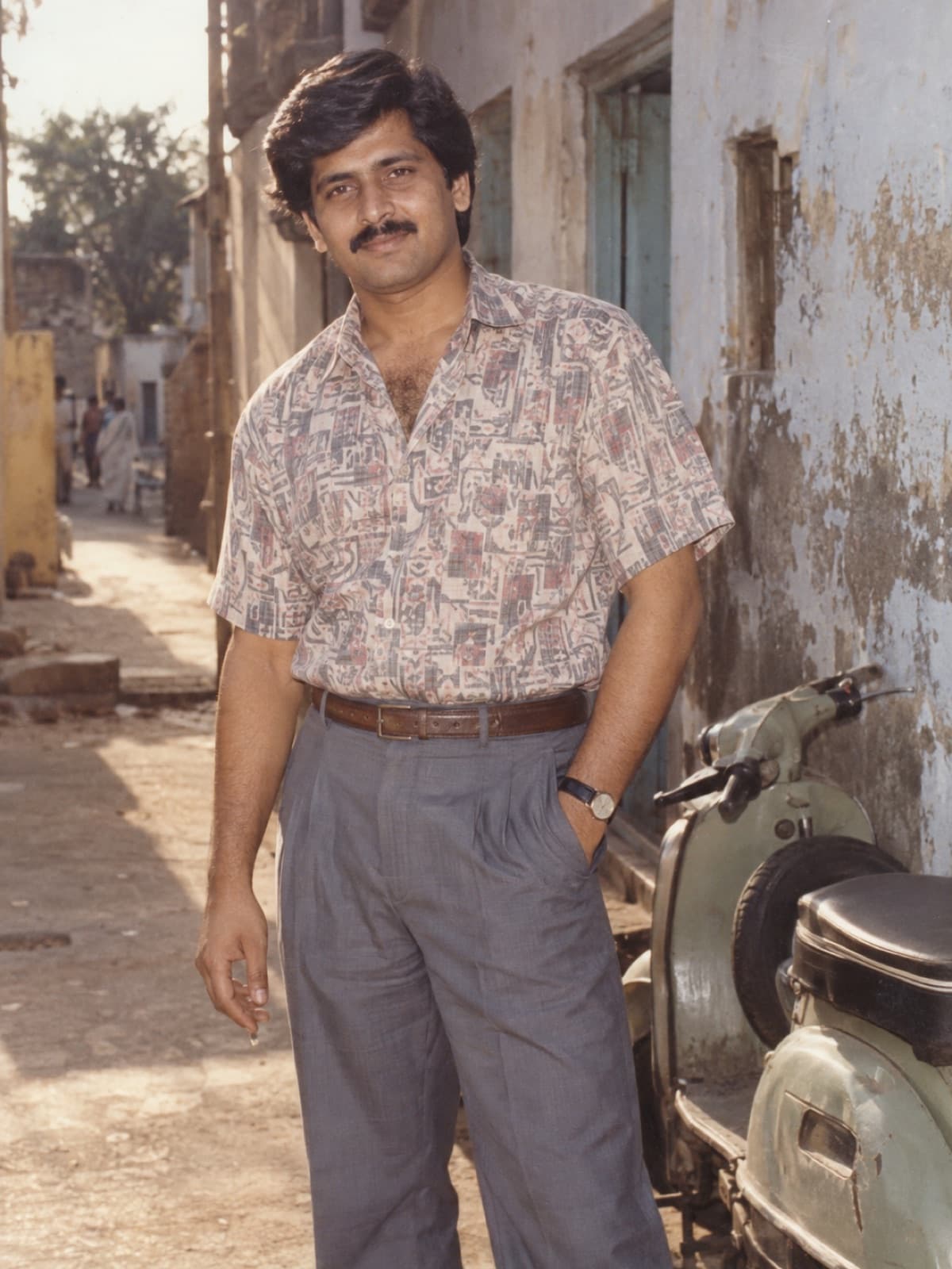AI-generated example of a South Asian man in a printed 1980s shirt and high-waisted trousers beside a vintage scooter on an old street